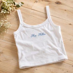 Op de afbeelding: Witte tanktop met dunne bandjes, met de naam "Mrs. Miller" in lichtblauw geborduurd. De top is gemaakt van een zachte stof en wordt gepresenteerd op een houten oppervlak, met bloemaccenten op de achtergrond.