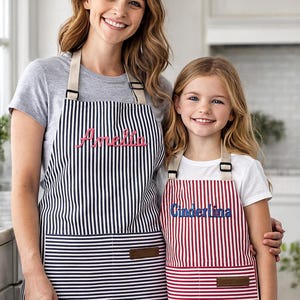 May include: Two aprons, one black and white striped, the other red and white striped. The black and white apron has the name "Amelia" embroidered in pink. The red and white apron has the name "Cinderlina" embroidered in blue.