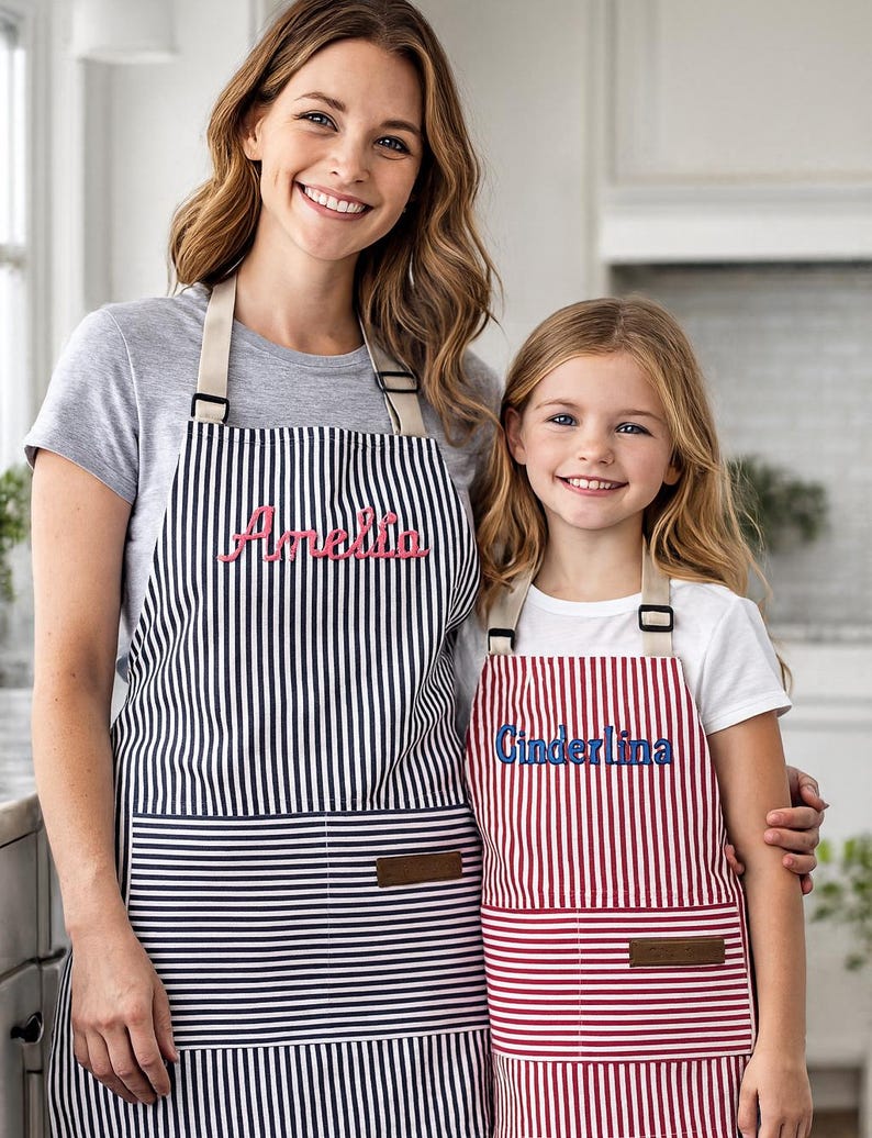 May include: Two aprons, one black and white striped, the other red and white striped. The black and white apron has the name "Amelia" embroidered in pink. The red and white apron has the name "Cinderlina" embroidered in blue.