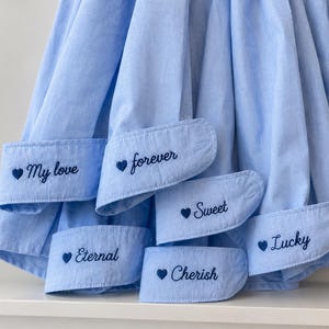 May include: Pale blue dress shirt with embroidered cuff details. The cuffs feature phrases such as "My love", "Forever", "Sweet", "Lucky", "Eternal", and "Cherish", each with a small heart. The shirt is made of a soft, light fabric.
