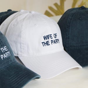 Könnte beinhalten: Eine Sammlung von Baseballkappen in Weiß und Marineblau. Eine weiße Kappe ist mit "WIFE OF THE PARTY" in Marineblau bestickt. Die marineblauen Kappen sind mit "THE PARTY" in Weiß bestickt.