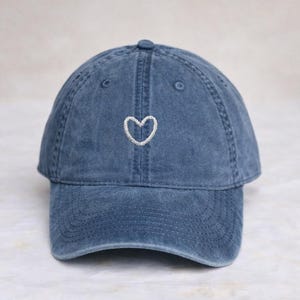 Peut inclure: Une casquette de baseball en jean bleu avec une finition délavée et usée. Le panneau avant présente un petit contour de cœur brodé en blanc, centré au-dessus de la visière. La casquette a une structure à six panneaux avec des œillets cousus.