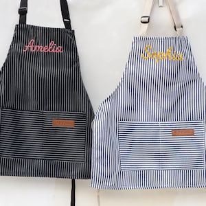 May include: Two striped aprons, one black and white, the other blue and white. The black apron has the name "Amelia" embroidered in pink. The blue apron has the name "Sophia" embroidered in yellow. Both have a front pocket and leather patch.