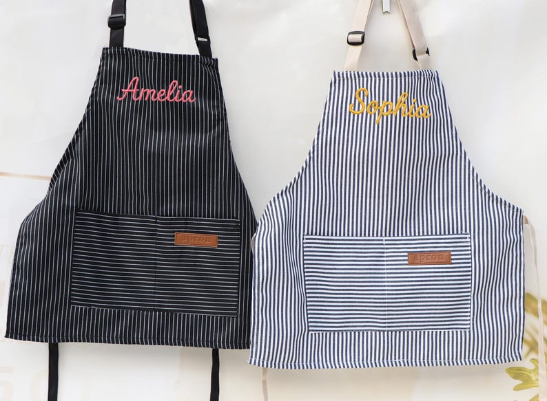 May include: Two striped aprons, one black and white, the other blue and white. The black apron has the name "Amelia" embroidered in pink. The blue apron has the name "Sophia" embroidered in yellow. Both have a front pocket and leather patch.