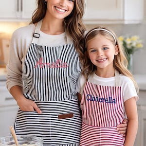 May include: Two aprons, one adult and one child-sized, with vertical stripes. The adult apron is navy and white with the name "Amelia" embroidered in pink. The child's apron is red and white with "Cinderella" in blue.