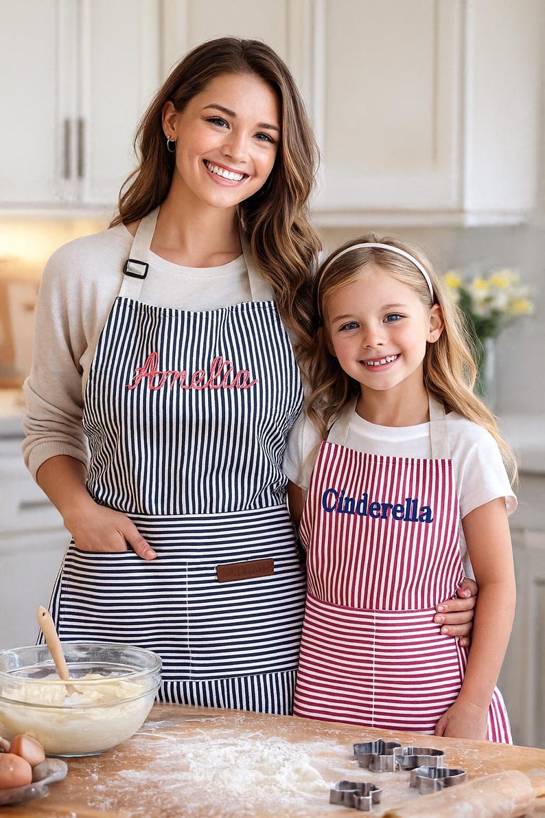 May include: Two aprons, one adult and one child-sized, with vertical stripes. The adult apron is navy and white with the name "Amelia" embroidered in pink. The child's apron is red and white with "Cinderella" in blue.