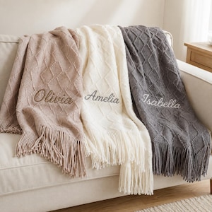 May include: Three knitted throw blankets in light beige, cream, and dark grey, each featuring a diamond-textured pattern and long fringe edges. The blankets are personalised with embroidered names: Olivia in brown, Amelia in grey, and Isabella in white.