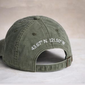 May include: An olive green baseball cap featuring white embroidered text on the back panel that reads 43.67° N, 121.50° W. The cap has a curved brim, a button on the crown, and an adjustable strap with a metal buckle closure at the back.