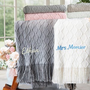 May include: A stack of soft blankets in various colors, including gray, pink, and white. The blankets have a diamond pattern and fringe. Two blankets are displayed, one gray with "Ethan" embroidered and one white with "Mrs Mercier".