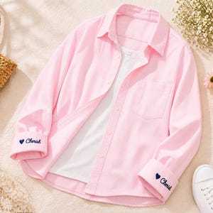 May include: A pale pink button-down shirt with a white undershirt. The shirt features a chest pocket and the word "Cherish" embroidered on the cuffs, accompanied by a blue heart. White trainers are also visible.