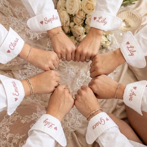 Puede incluir: Un grupo de personas con camisas blancas abotonadas con pu&ntilde;os personalizados. Cada pu&ntilde;o est&aacute; bordado con un nombre en escritura rosa y un peque&ntilde;o coraz&oacute;n. Las manos est&aacute;n unidas en c&iacute;rculo.