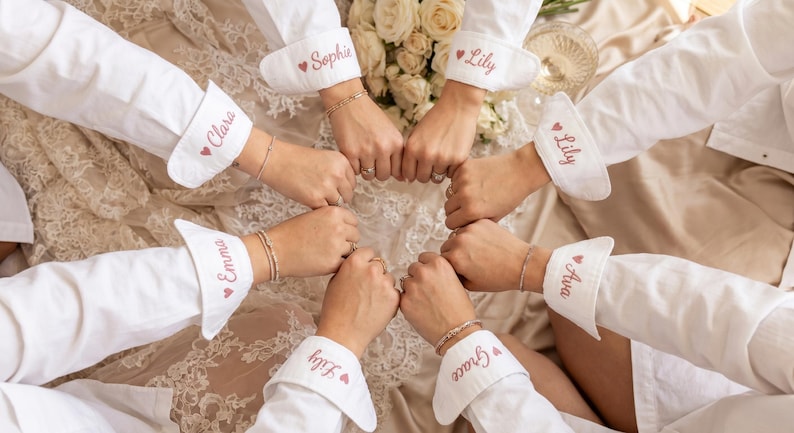 Puede incluir: Un grupo de personas con camisas blancas abotonadas con pu&ntilde;os personalizados. Cada pu&ntilde;o est&aacute; bordado con un nombre en escritura rosa y un peque&ntilde;o coraz&oacute;n. Las manos est&aacute;n unidas en c&iacute;rculo.