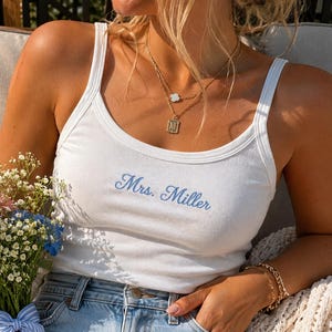 Può includere: Canotta bianca a coste con il nome "Mrs. Miller" ricamato in corsivo blu chiaro. Il top è abbinato a jeans blu. Un bouquet di fiori e collane d'oro completano il look.