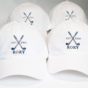 Puede incluir: Cuatro gorras de béisbol blancas con diseño de palos de golf bordados. El diseño incluye palos de golf cruzados, el texto "EST 2025" y el nombre "RORY" en hilo azul y negro. Las gorras están dispuestas sobre una superficie blanca.