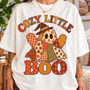 May include: A white short-sleeve t-shirt featuring a graphic of a ghost wearing a witch hat. The ghost is composed of various autumn-themed fabric patterns including plaid, leaves, and acorns. The text COZY LITTLE BOO is printed in orange.