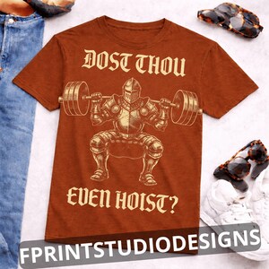 K&ouml;nnte beinhalten: Rostfarbenes T-Shirt mit einem Ritter, der Gewichte hebt, mit dem Text "DOST THOU EVEN HOIST?" in einer mittelalterlichen Schriftart. Das Design beinhaltet eine detaillierte Illustration des Ritters in R&uuml;stung. Teil einer Bekleidungskollektion.