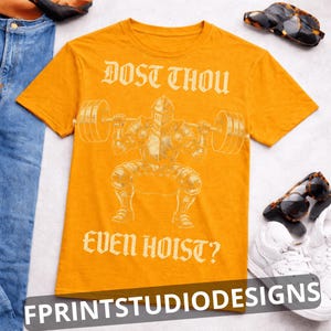 K&ouml;nnte beinhalten: Senfgelbes T-Shirt mit einer Grafik eines Ritters, der Gewichte hebt, und dem Text "DOST THOU EVEN HOIST?". Das Shirt hat auch den Text "FPRINTSTUDIODESIGNS" unten. Sonnenbrillen und wei&szlig;e Turnschuhe sind ebenfalls sichtbar.