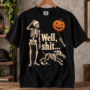 May include: A black short-sleeved t-shirt featuring a graphic of a skeleton scratching its head, a skeleton dog running with a hand in its mouth, and an orange jack-o'-lantern balloon. The text "Well, shit..." is printed in cream-coloured letters.