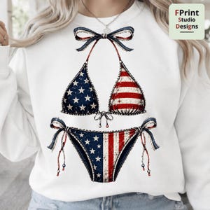 Peut inclure: Un sweat-shirt blanc à manches longues avec une impression graphique d'un bikini au motif de drapeau américain vieilli. Le haut du bikini présente un nœud au centre et le bas du bikini est doté de liens latéraux. Le motif affiche des tons rouge, blanc et bleu.