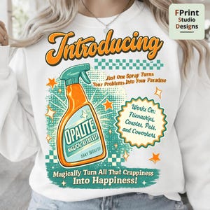 Pode incluir: Camisola branca com um gráfico com um borrifador laranja com a etiqueta "Opalite" com o texto "Transforme magicamente toda essa porcaria em felicidade!" e "Apenas um spray transforma seus problemas em seu paraíso."