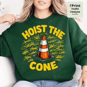 May include: A dark green sweatshirt with the words "HOIST THE CONE" in yellow, surrounding an orange and white traffic cone graphic. The design includes yellow signatures. The sweatshirt is a crew neck style.