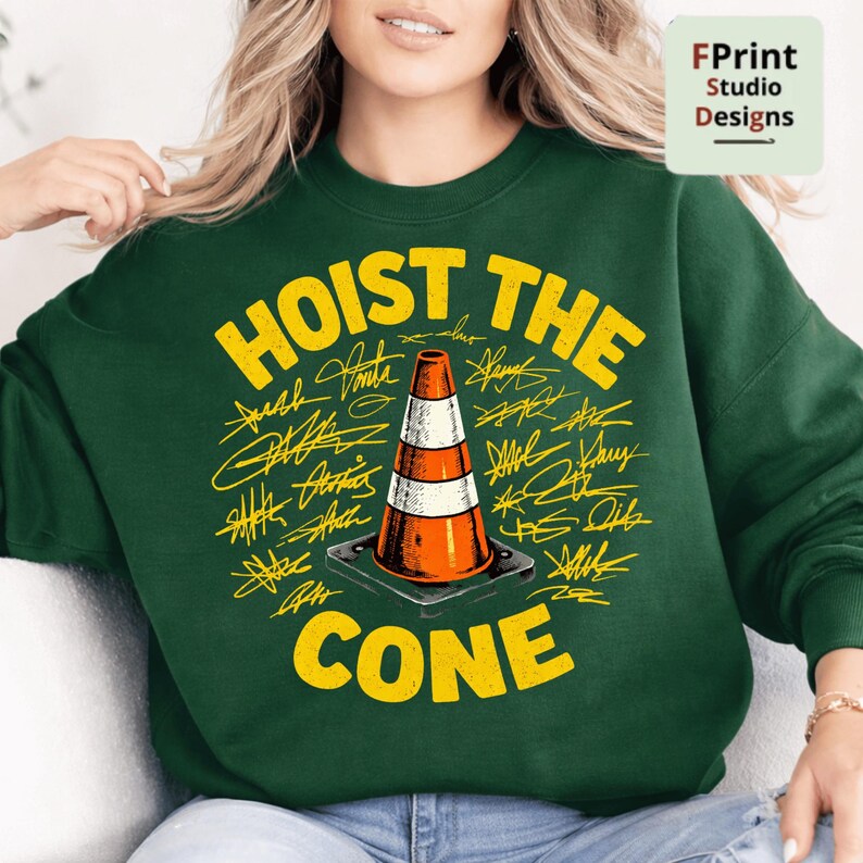 May include: A dark green sweatshirt with the words "HOIST THE CONE" in yellow, surrounding an orange and white traffic cone graphic. The design includes yellow signatures. The sweatshirt is a crew neck style.