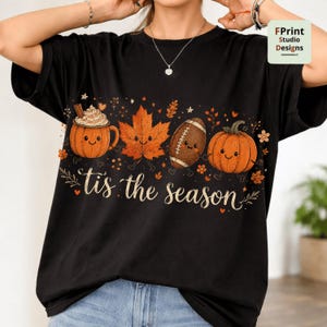 以下が含まれることがあります： 白い筆記体で「tis the season」という文字がプリントされた黒の半袖Tシャツ。ホイップクリームが乗った笑顔のパンプキンマグカップ、カエデの葉、アメリカンフットボール、そしてパンプキンが描かれており、それぞれに顔がデザインされています。