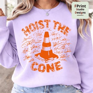 May include: A light purple sweatshirt with the words "HOIST THE CONE" in orange, along with an orange traffic cone graphic. The design includes what appears to be signatures around the cone. The sweatshirt is a casual style.