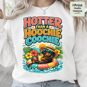 May include: A white sweatshirt featuring a graphic of a dachshund wearing a straw hat and blue sunglasses, floating in a colorful pool ring while holding a drink. The text reads HOTTER THAN A HOOCHIE COOCHIE in orange, yellow, and blue letters.