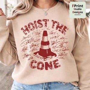 May include: Beige sweatshirt with a distressed red graphic that reads "HOIST THE CONE" above a traffic cone illustration. The design includes signatures around the cone. The sweatshirt has long sleeves.