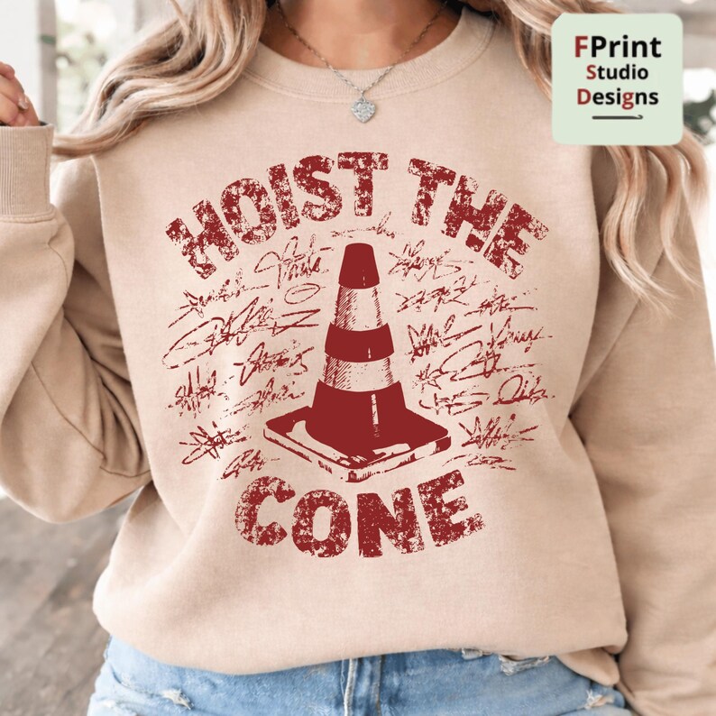 May include: Beige sweatshirt with a distressed red graphic that reads "HOIST THE CONE" above a traffic cone illustration. The design includes signatures around the cone. The sweatshirt has long sleeves.