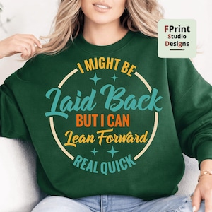 Puede incluir: Sudadera verde oscuro con el texto "I might be laid back but I can lean forward real quick" en un diseño circular. El texto es en color turquesa, naranja y crema. La sudadera tiene mangas largas.