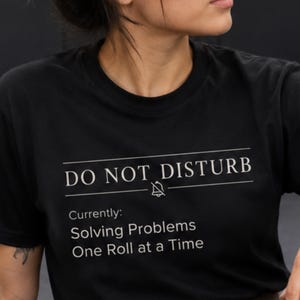 Op de afbeelding: Een zwart t-shirt met korte mouwen en witte tekst met de tekst DO NOT DISTURB met een doorgestreept bel-icoon, gevolgd door de tekst Currently: Solving Problems One Roll at a Time. Het ontwerp is gecentreerd op de borst van het kledingstuk.