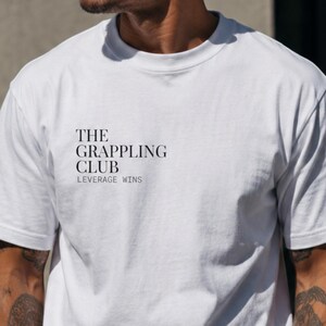 Peut inclure: Un t-shirt blanc à manches courtes avec du texte noir sur la poitrine gauche. Le texte indique THE GRAPPLING CLUB dans une police serif grasse, avec les mots LEVERAGE WINS imprimés dans une police sans-serif plus fine juste en dessous.