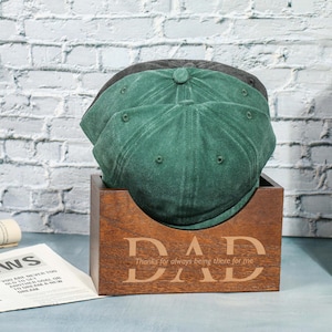 May include: A wooden box engraved with the word "DAD" and the phrase "Thanks for always being there for me", holding three hats. The hats are green and grey. The box is on a table.