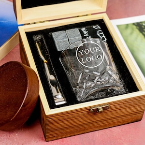 Op de afbeelding: Houten geschenkdoos met een whiskyglas met een ruitpatroon en de tekst "YOUR LOGO", whisky stenen en een metalen ijstang. De doos is open en onthult de inhoud op een zwarte fluwelen voering.