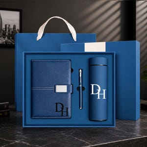 May include: A blue gift set containing a notebook with a silver clasp and the initials DH, a slim blue pen, and a cylindrical blue vacuum flask featuring the white initials DH. The items are presented in a matching blue box and gift bag.