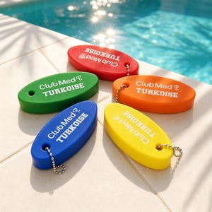May include: Five oval-shaped floating keychains in red, green, orange, blue, and yellow. Each keychain features the white text Club Med TURKOISE and a trident logo. A small hole at one end of each keychain is attached to a silver metal ball chain.