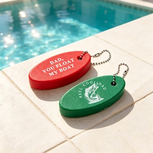 May include: Two oval-shaped foam keychains with metal ball chains. The red keychain features the white text "DAD, YOU FLOAT MY BOAT." The green keychain displays a white graphic of a fish jumping out of water with the text "REEL COOL DAD" printed around it.