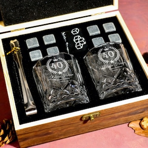 Op de afbeelding: Houten geschenkdoos met twee gegraveerde whiskyglazen, ijsblokjes en een tang. De glazen zijn geëtst met een "40"-ontwerp en gepersonaliseerde tekst. De set bevat grijze stenen ijsblokjes en een goudkleurige ijstang.