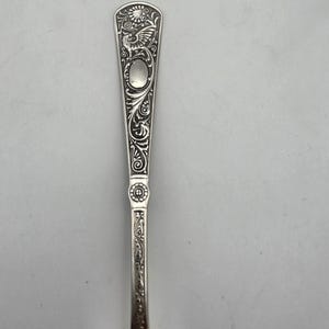 May include: A silver-toned metal spoon handle featuring an intricate embossed design. The handle displays a winged creature at the top, a smooth oval medallion, scrolling floral patterns, and a small circular floral motif near the center.