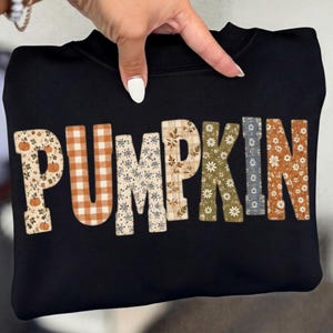 Peut inclure: Un sweat-shirt noir avec le mot PUMPKIN écrit en grandes lettres majuscules. Chaque lettre présente un motif distinct, incluant des citrouilles orange sur fond crème, un vichy orange et blanc, des fleurs bleues, un tartan beige et des marguerites.