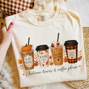 Könnte beinhalten: Ein cremefarbenes T-Shirt mit einer Grafik von vier Kaffeebechern in herbstlichen Designs, inklusive der Schriftzüge hello FALL, AUTUMN vibes, SWEATER WEATHER und Pumpkin SPICE sowie dem Satz Autumn leaves & coffee please darunter.