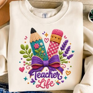 Könnte beinhalten: Ein cremefarbenes Sweatshirt mit Rundhalsausschnitt und einer Grafik aus einem Bleistift und einem Radiergummi, die mit einer violetten Schleife zusammengebunden sind. Das Design enthält florale Akzente, kleine Herzen und den Text "Teacher Life" in violetter Schreibschrift.
