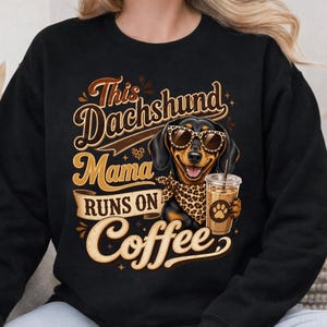 May include: A black sweatshirt featuring a graphic of a dachshund wearing leopard print sunglasses and a matching bandana, holding a plastic cup of iced coffee. The text reads: This Dachshund Mama RUNS ON Coffee, styled in brown and cream lettering.