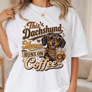 May include: A white short-sleeve t-shirt featuring a graphic of a black and tan dachshund wearing leopard print sunglasses and a matching bandana. The dog holds a plastic cup of iced coffee. The text reads: This Dachshund Mama Runs on Coffee.