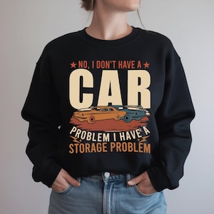May include: A black crewneck sweatshirt featuring a graphic print with the text "NO, I DON'T HAVE A CAR PROBLEM I HAVE A STORAGE PROBLEM" in beige and orange. The design includes illustrations of two vintage cars in orange and blue tones.