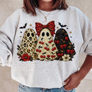 May include: A light gray crewneck sweatshirt featuring a graphic of three ghosts. The left ghost has a leopard print, the middle ghost has a red bow and cherry pattern, and the right ghost is black with cherry and skull motifs, accented by bats and stars.