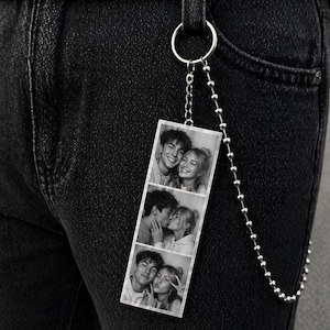 Puede incluir: Un llavero con una tira de fotos en blanco y negro que muestra tres imágenes de una pareja. El llavero está sujeto a unos vaqueros negros con una cadena y un anillo plateados.