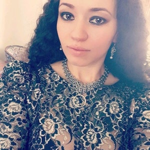 May include: A woman wearing a black lace top with a silver chain necklace and silver earrings. The necklace has a large silver ring pendant.